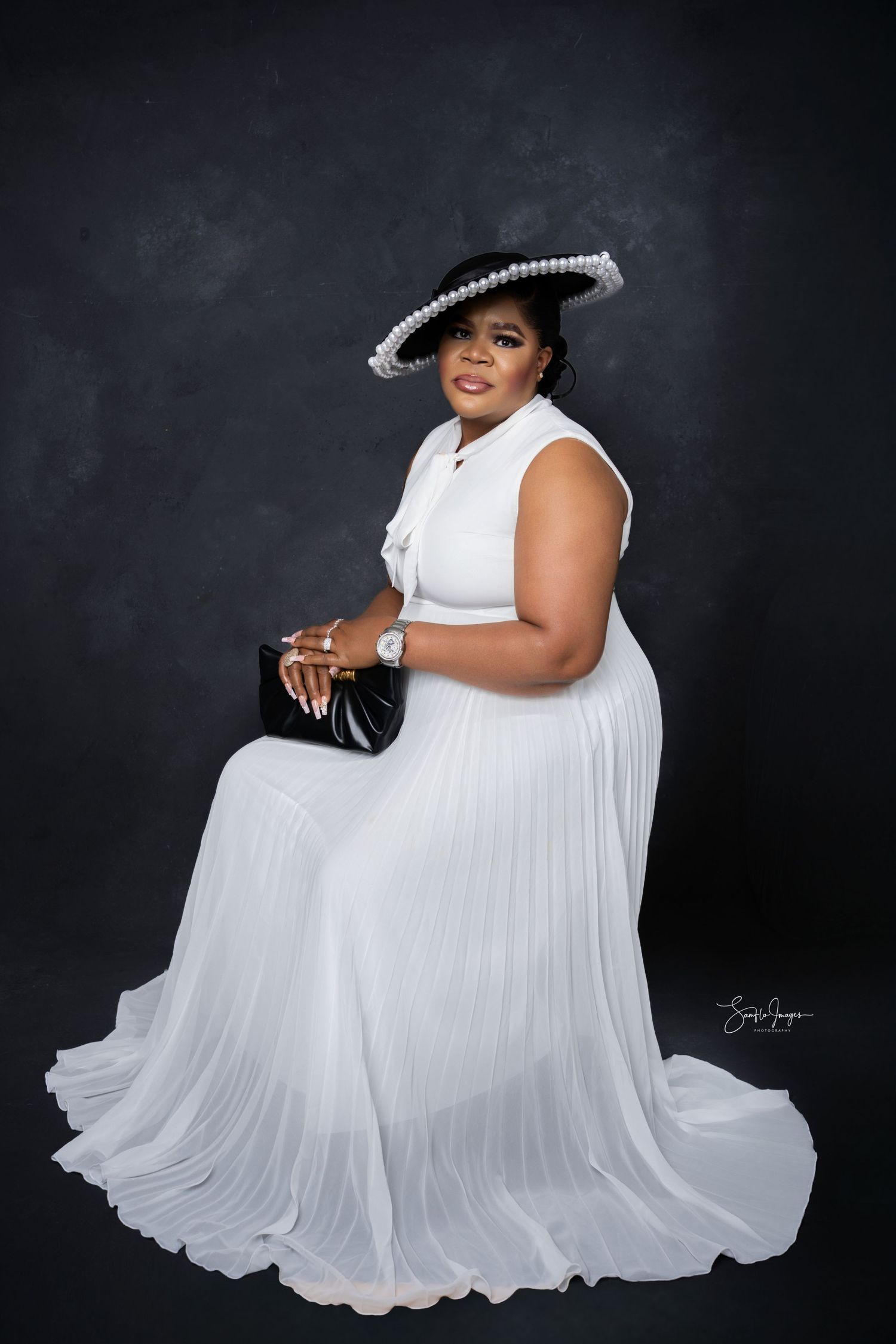Woman in white pleated gown and black pearl-trimmed hat poses elegantly for a formal portrait photo.