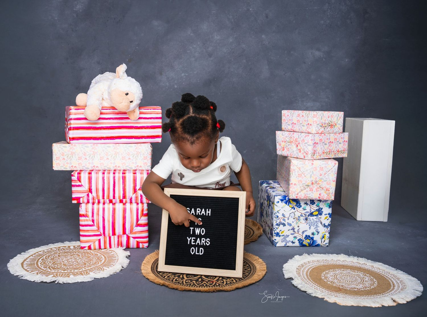 Toddler girl sits with gift boxes and a letter board sign reading Sarah Two Years Old for a birthday photo.