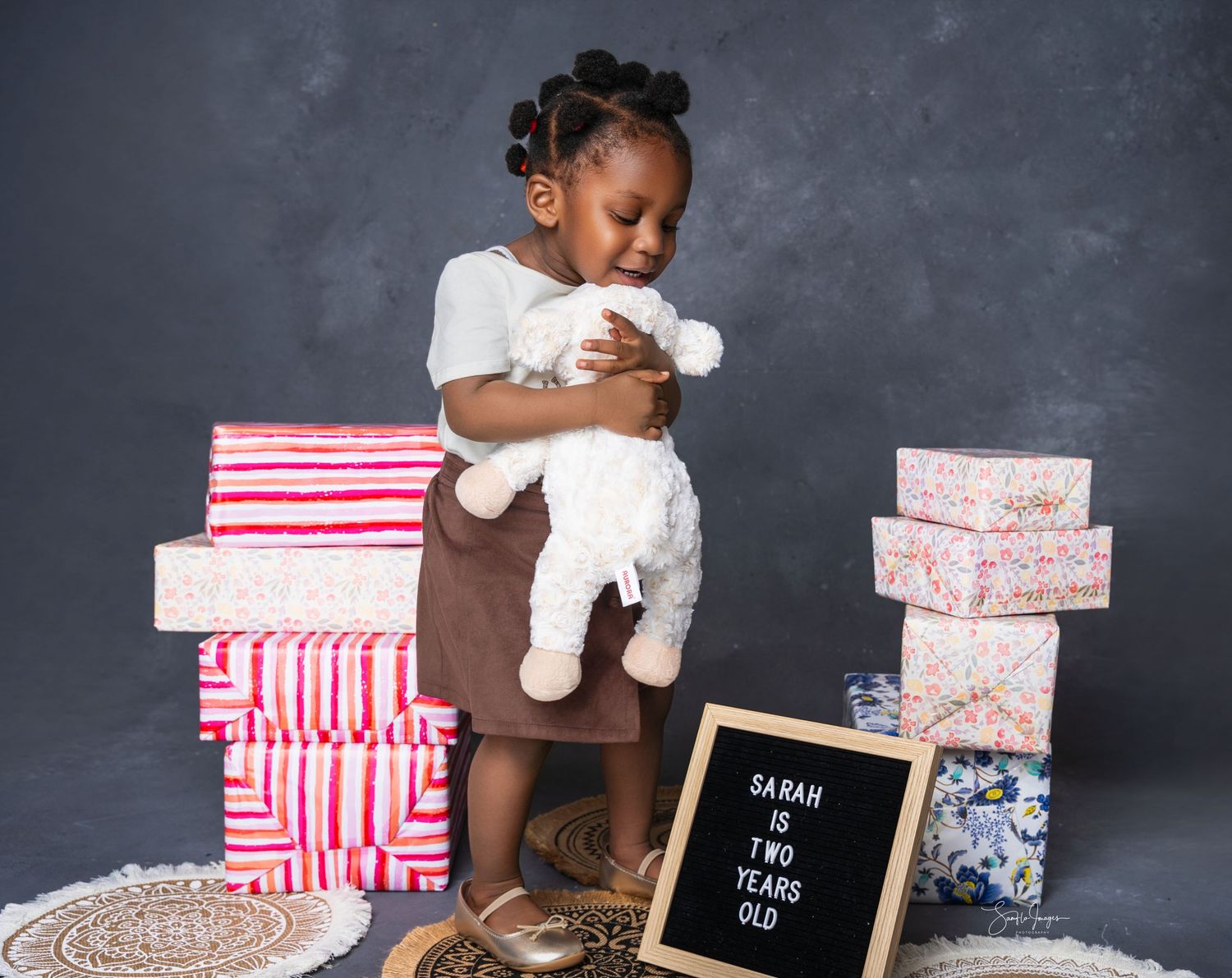 Toddler girl Sarah holds a stuffed animal surrounded by gift boxes, celebrating her second birthday.