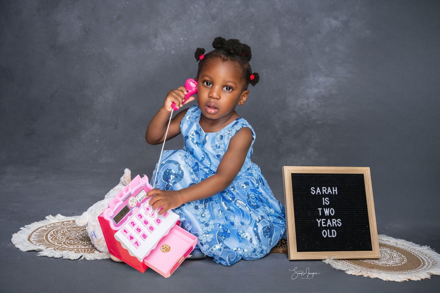 Toddler girl in blue dress holds pink toy phone next to a letterboard sign reading Sarah Is Two Years Old.