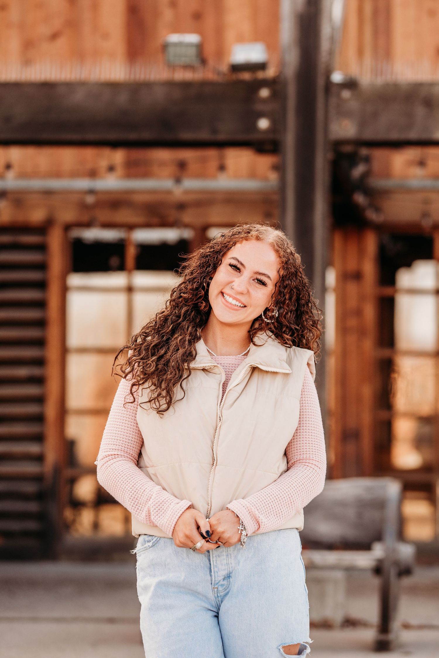 A person wearing a cream vest and pink sweater stands smiling in front of a wooden building.