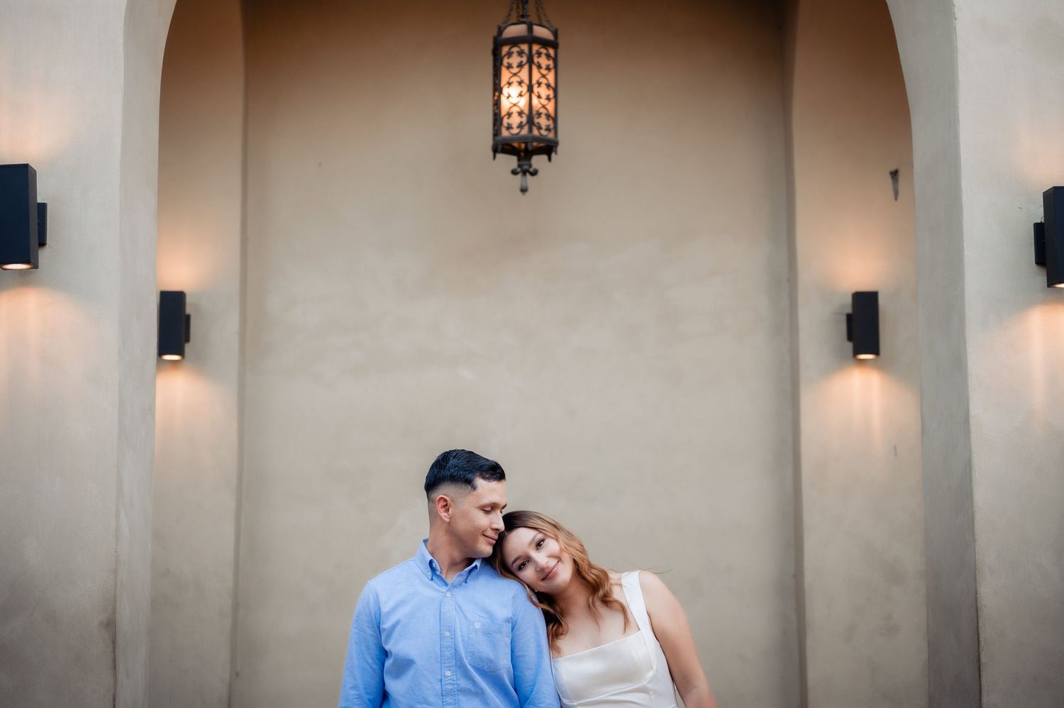Couple embraces under arched walkway with warm lighting and lantern fixture against beige stucco wall.