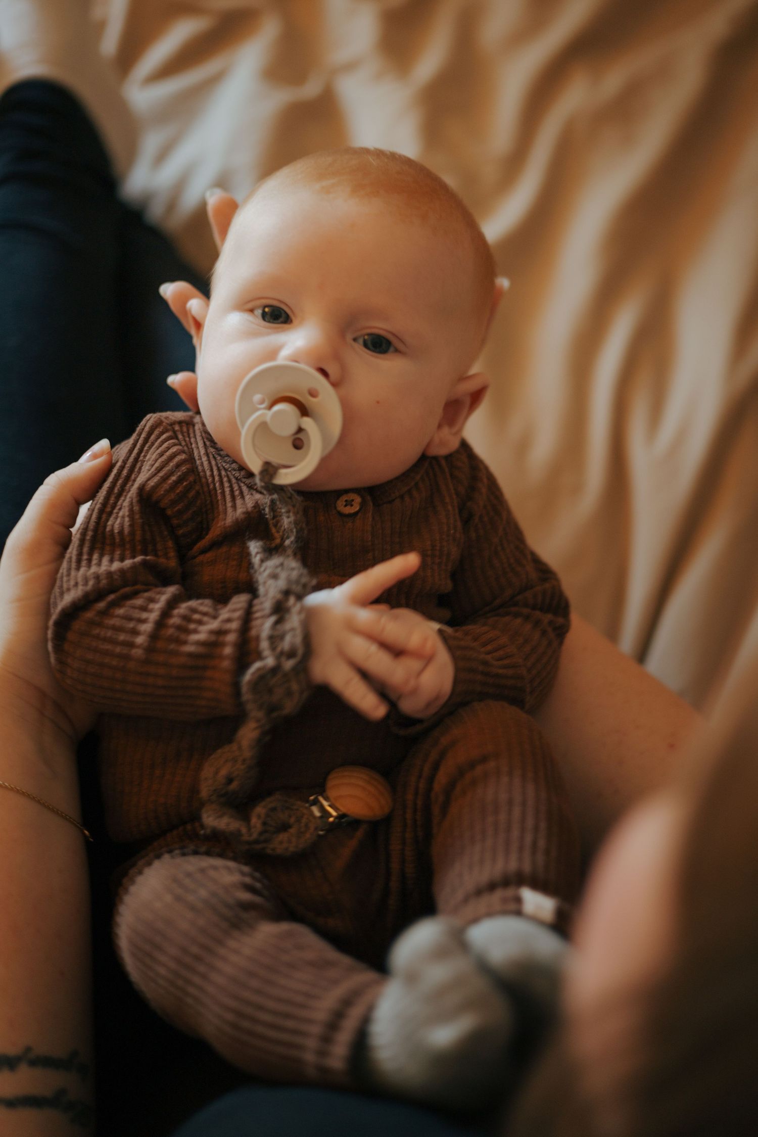 Baby in brown striped onesie with pacifier lying on beige bedding.
