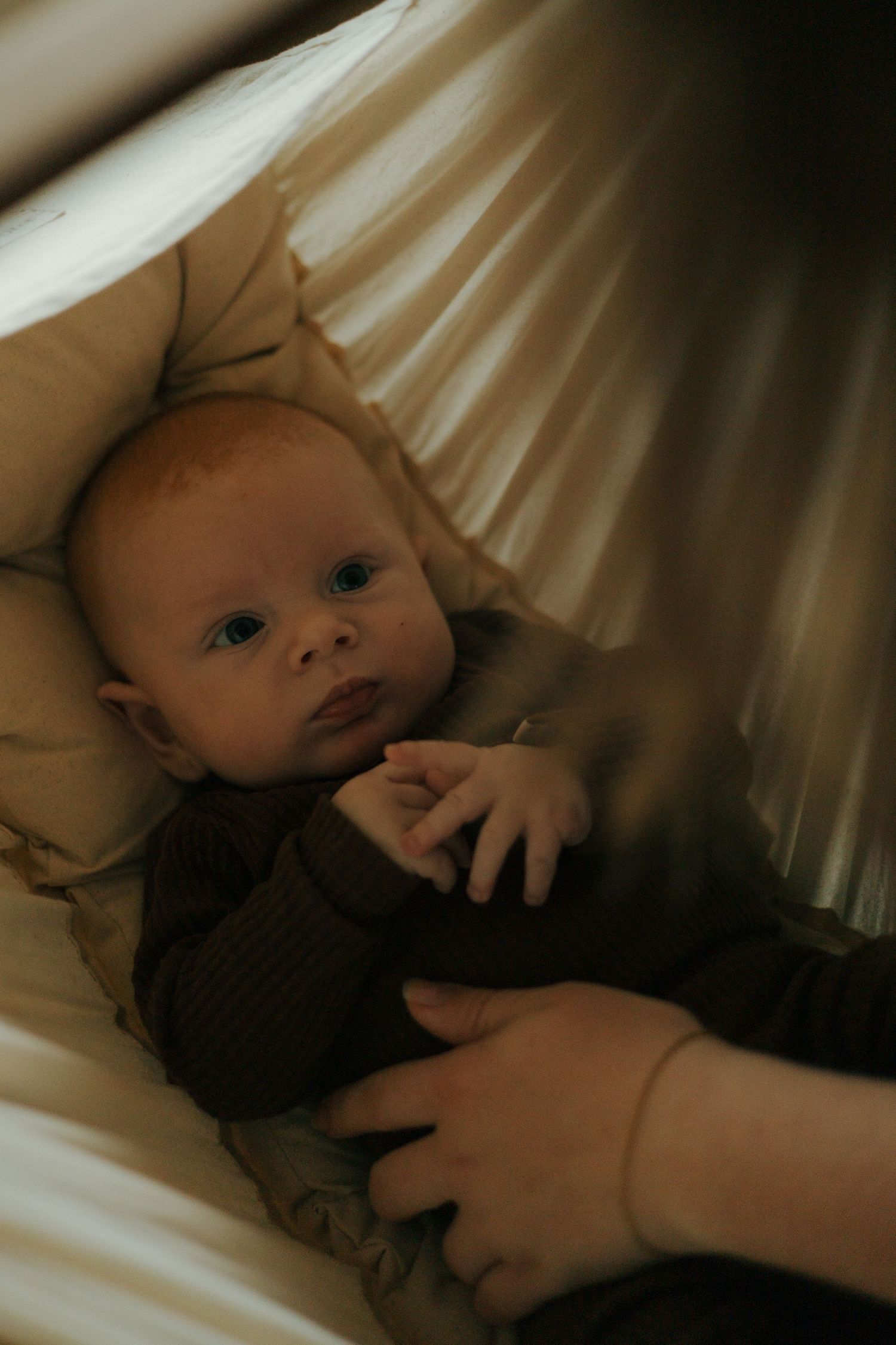 Baby in brown sweater resting on white bedding with gentle lighting.