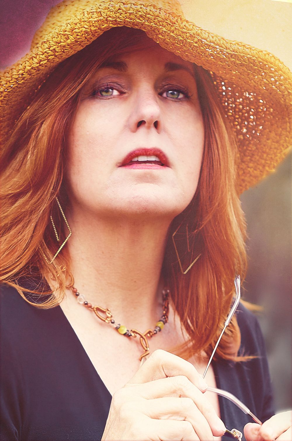 Portrait of someone wearing a straw hat and diamond earrings with red hair against a warm-toned background.