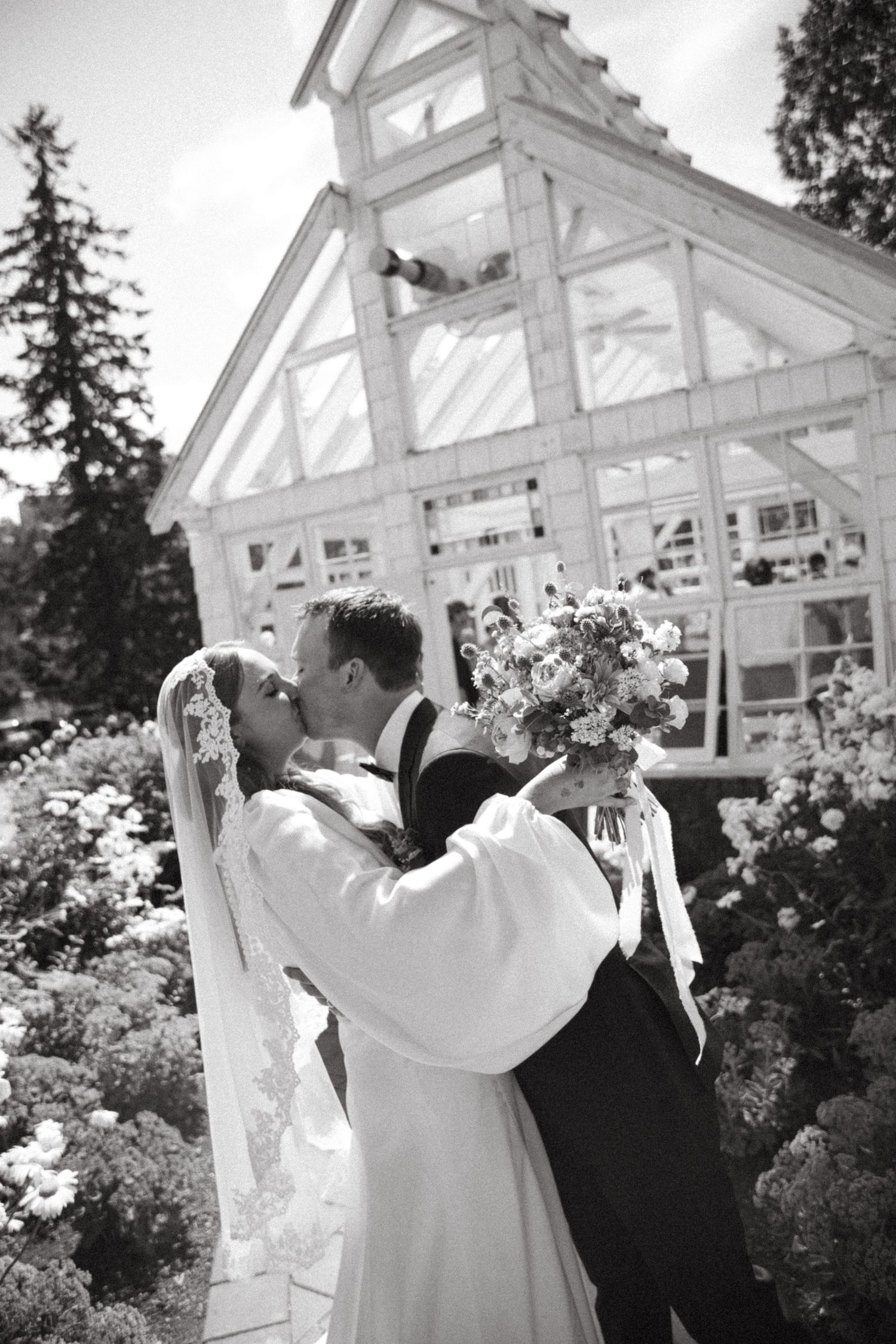 Black and white photo of a bride and groom kissing in front of a white greenhouse surrounded by blooming garden flowers.