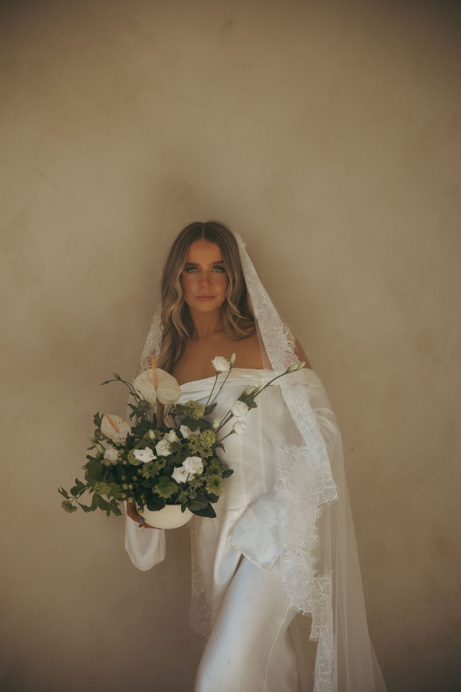 Elegant bride in a white satin gown with lace veil holds a green and white bouquet while posing against a neutral wall.