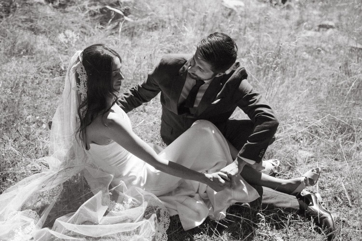 Black and white photo of a bride and groom sitting together in a grassy field, holding hands and sharing an intimate moment.