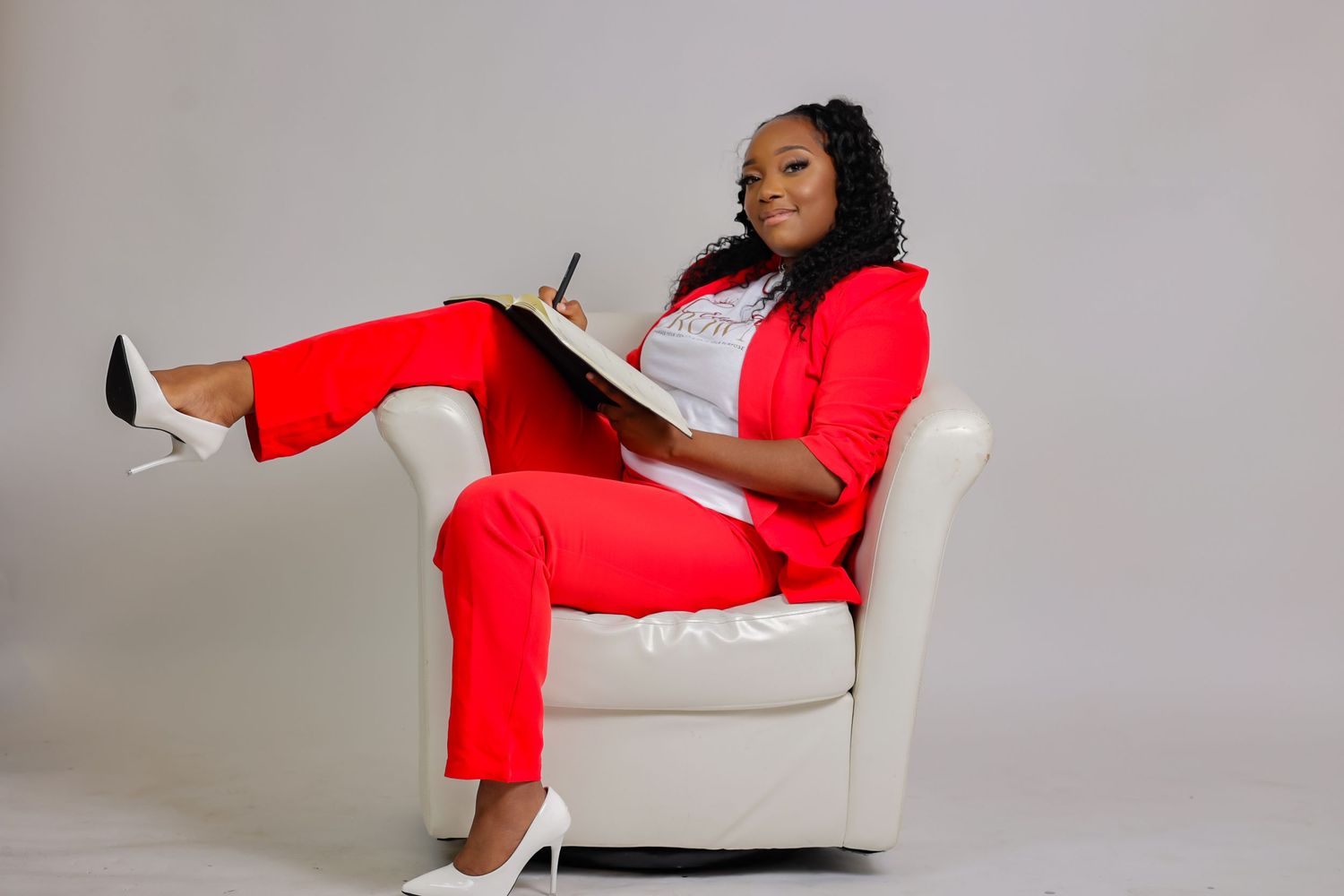 Confident woman in red suit sitting in white chair writing notes in a professional portrait.