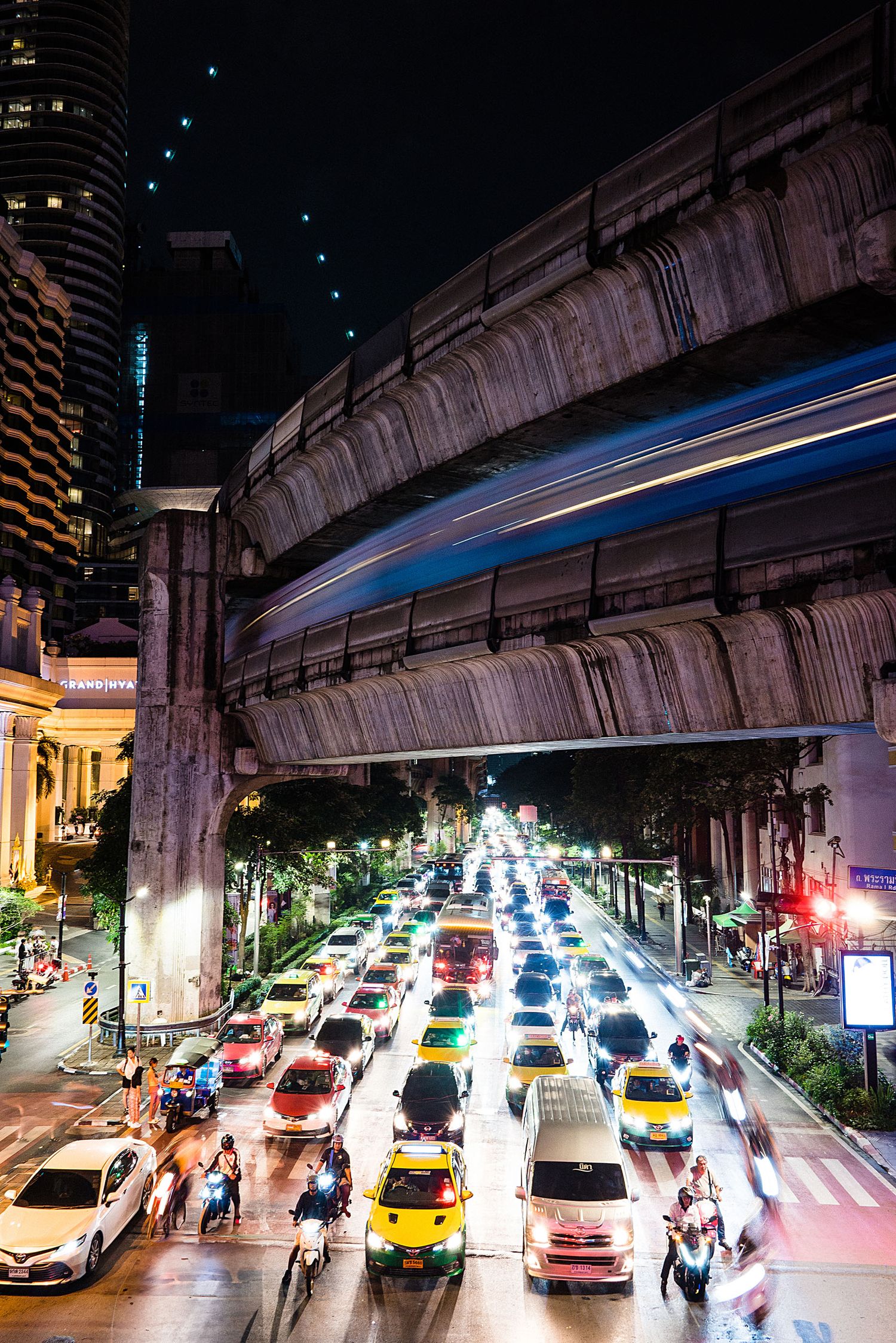 Busy nighttime street in Bangkok with colorful taxis and cars under an elevated train track with city lights.
