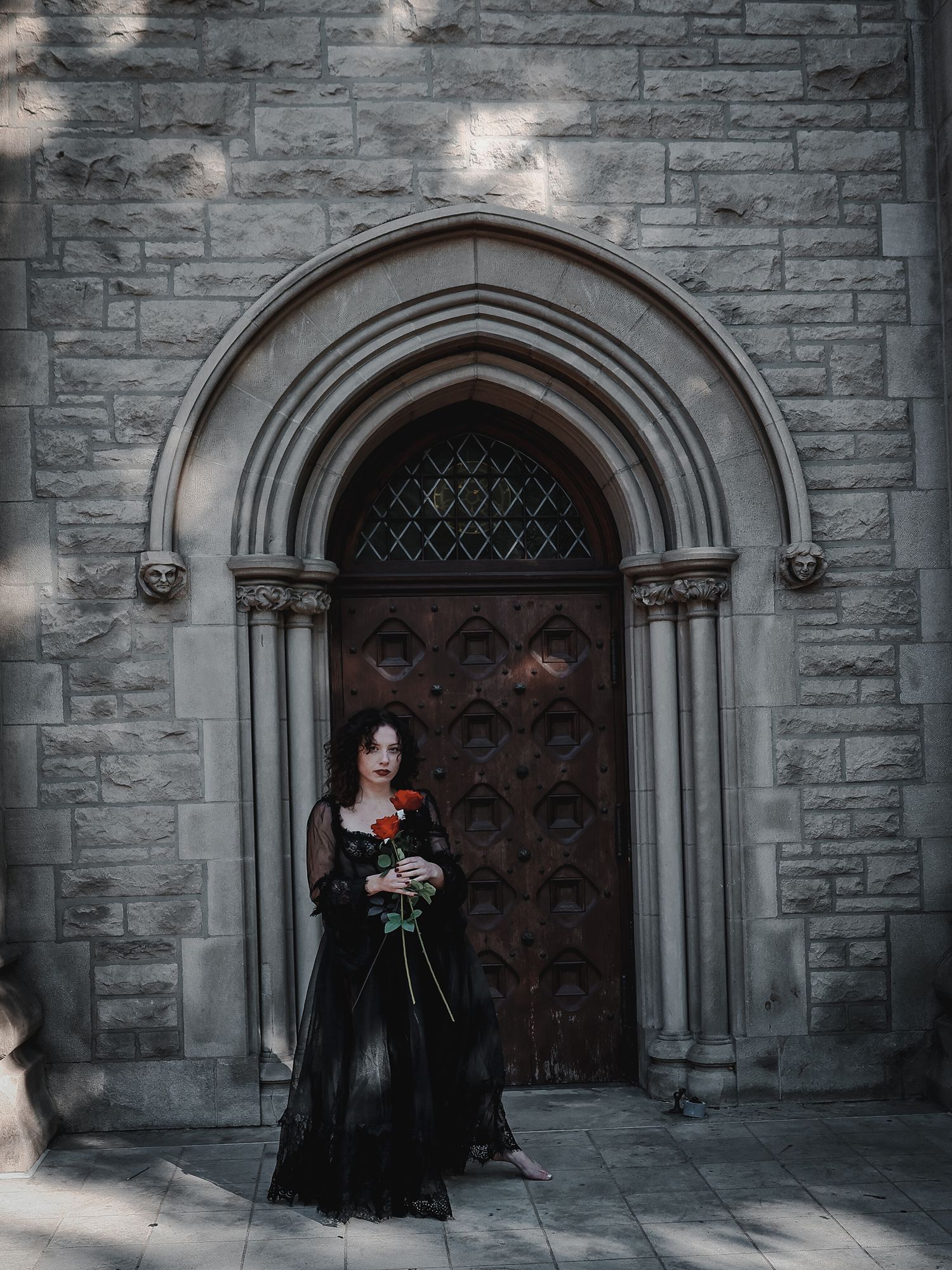 A gothic-style church doorway frames a figure in black holding a red rose against medieval stonework architecture.