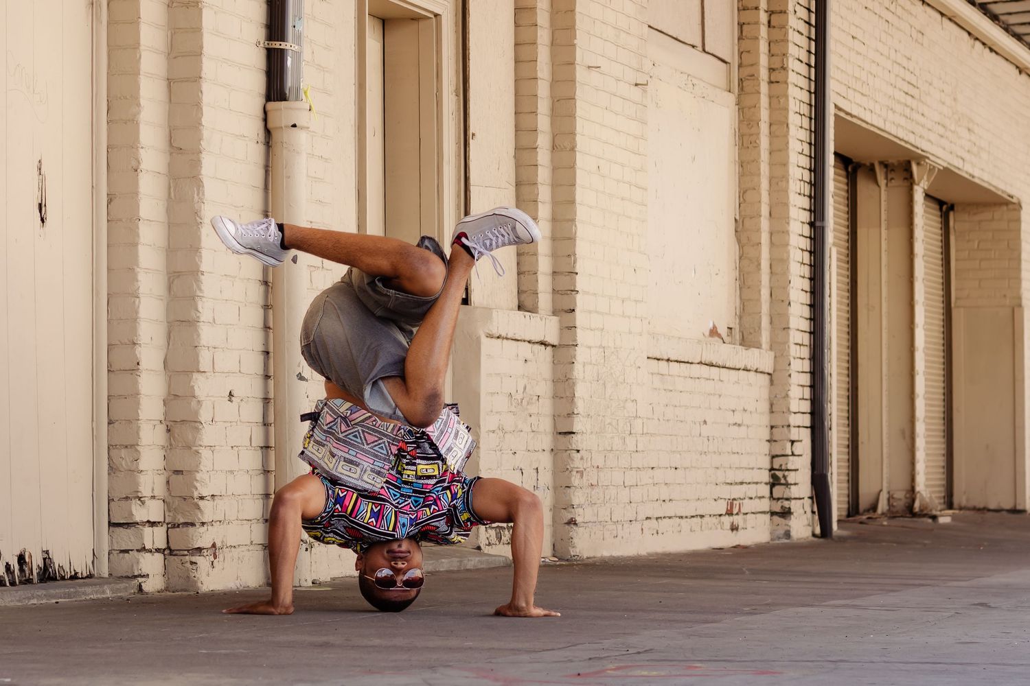 A breakdancer performs a headstand against a white brick wall in an urban alley.
