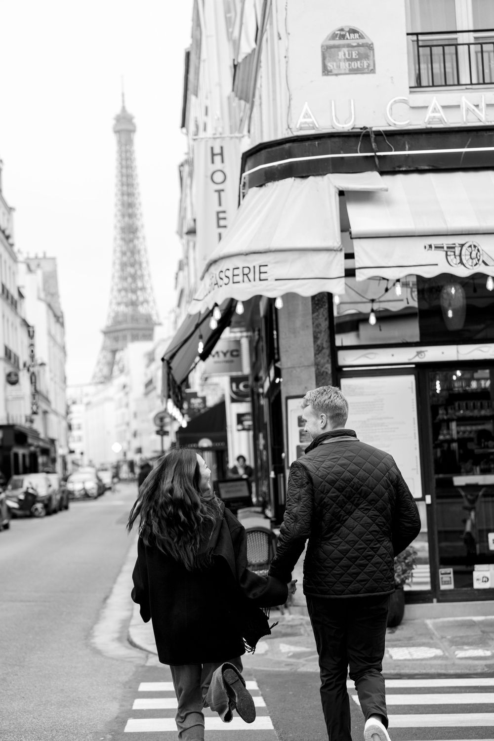 A black and white photo of a couple walking down a Paris street with the Eiffel Tower visible in the background.