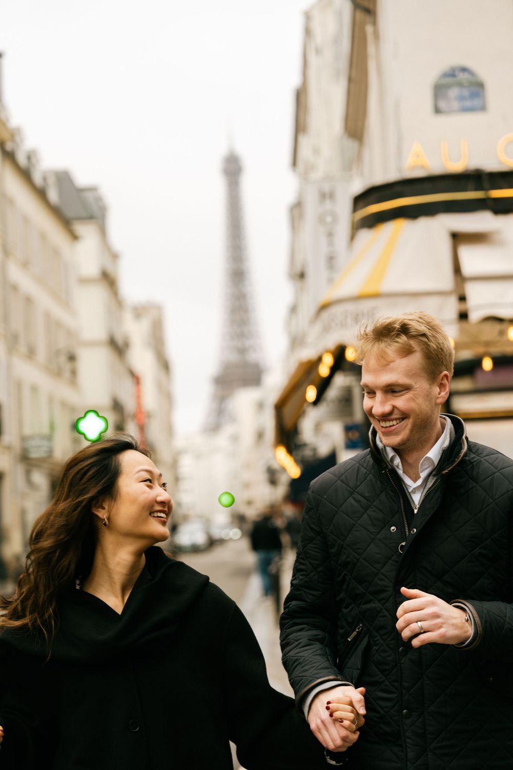 A couple shares a joyful moment while walking on a Parisian street with the Eiffel Tower visible in the background.