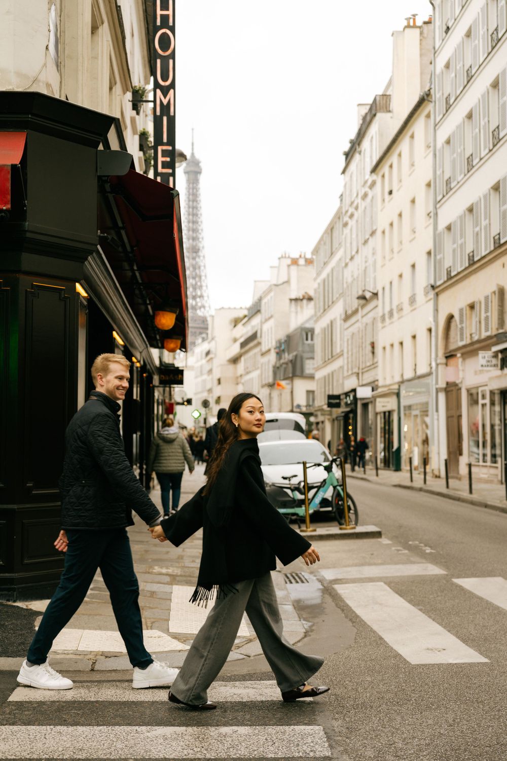 Couple walking hand in hand across a crosswalk on a charming Parisian street with the Eiffel Tower visible in the distance.