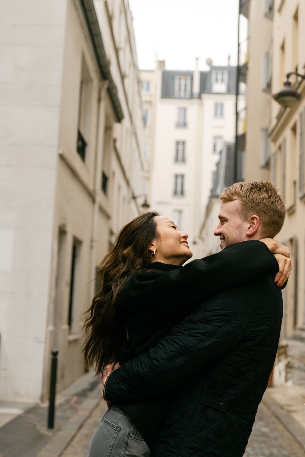 A romantic couple embraces and laughs together on a charming Parisian street with classic architecture in the background.