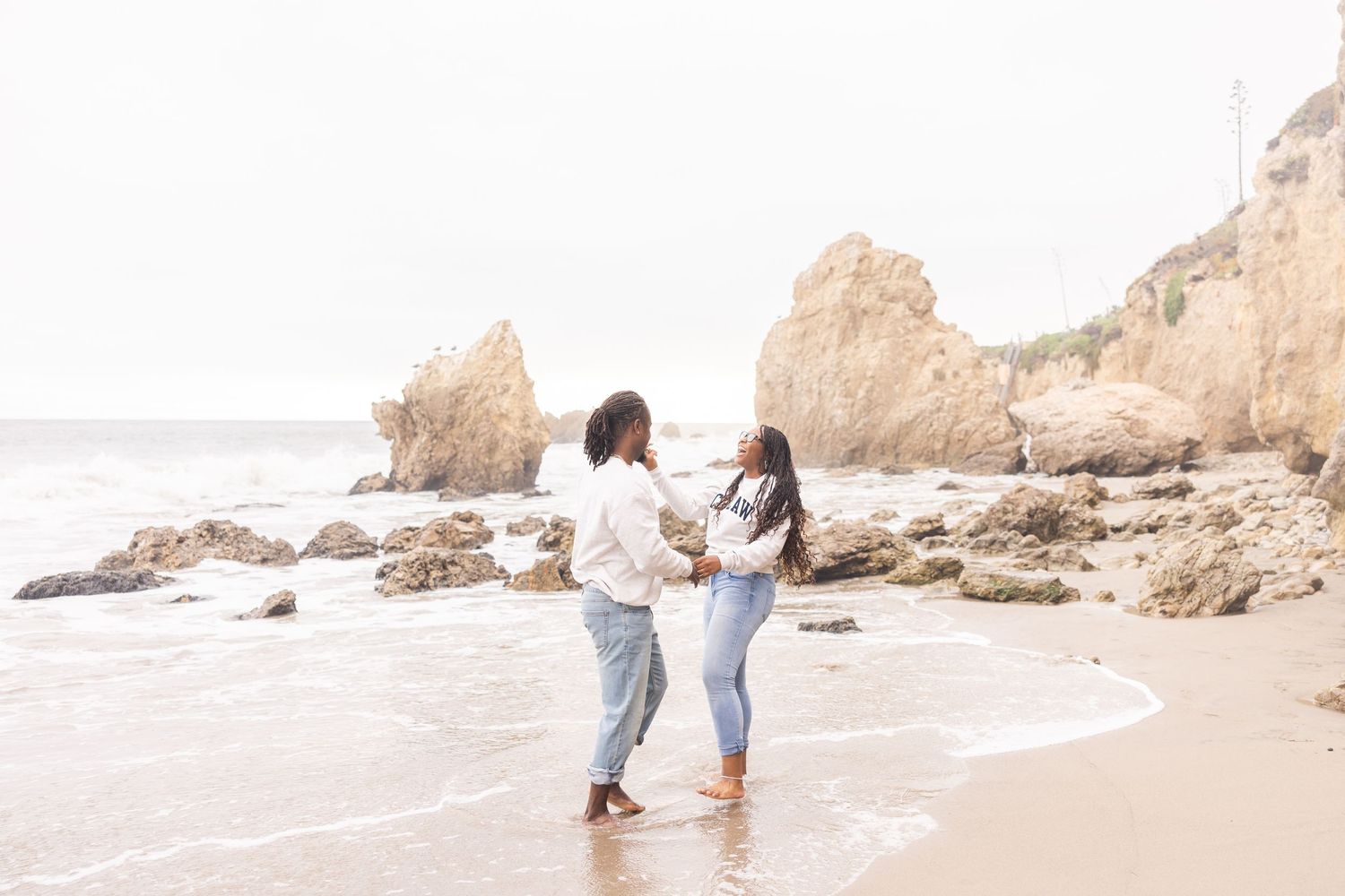 A couple hold hands while standing on a serene beach with rocky coastal cliffs and waves in the background.