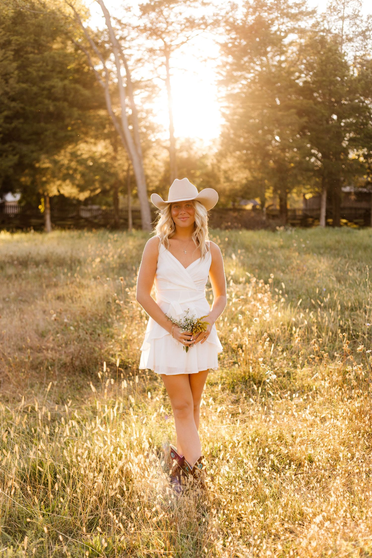A white sundress and cowboy hat glow in golden sunlight against a rustic outdoor setting at sunset.
