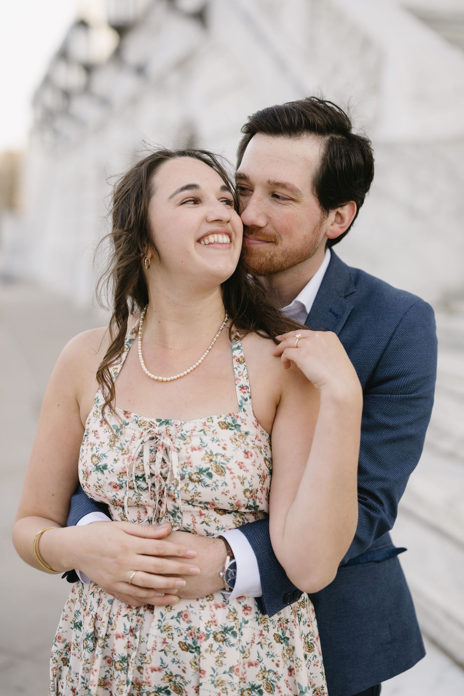 A romantic couple embraces outdoors, with her in a floral dress and him in a navy suit.