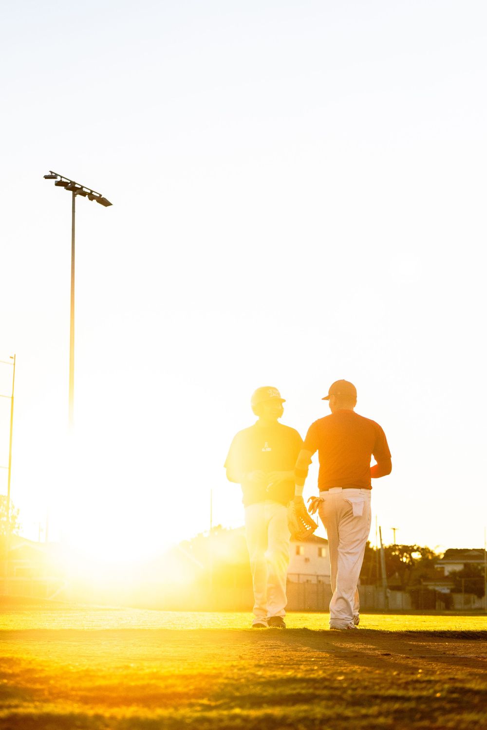 Baseball players practicing on field at golden hour with dramatic sunlight casting long shadows across the grass.