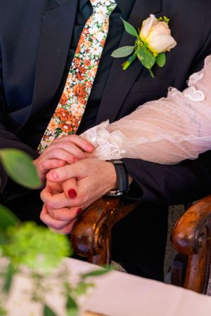 Close-up of clasped hands at wedding ceremony showing floral tie, pink lace dress sleeve and peach rose boutonniere.