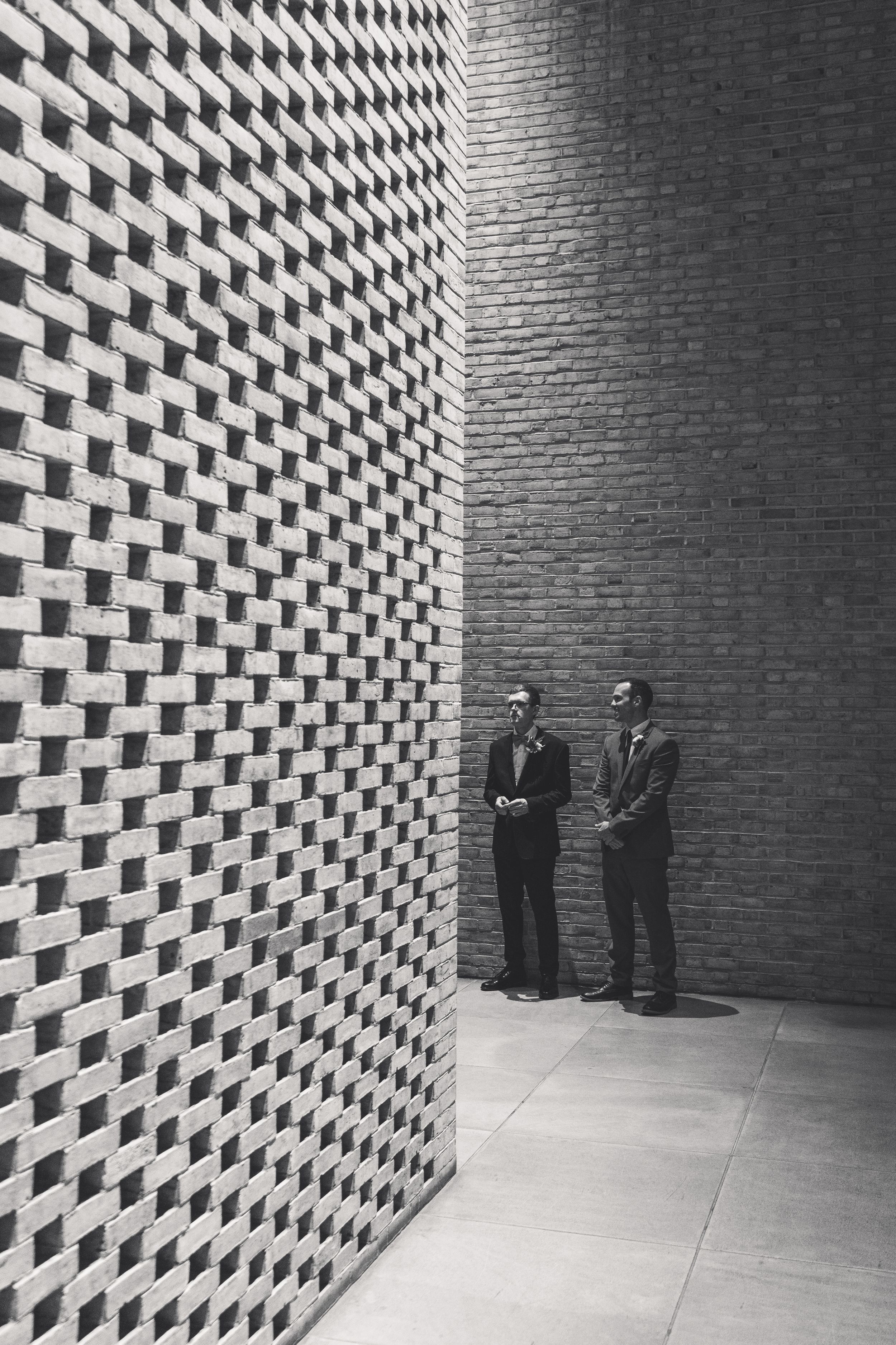 Two figures stand beside a geometric patterned wall creating stark contrast against brick in black and white photo.