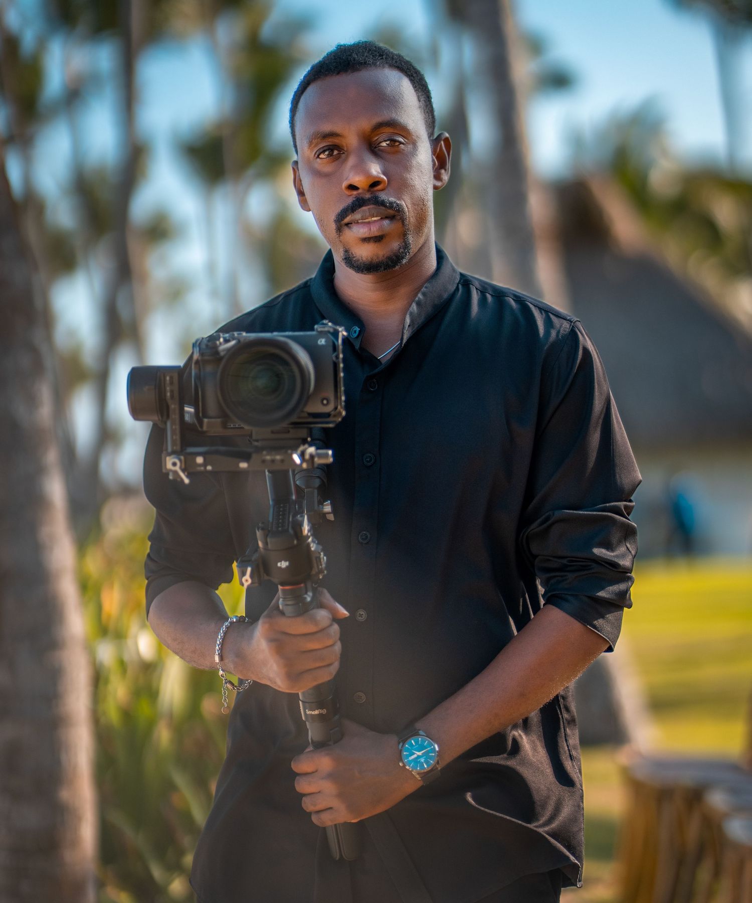 A photographer wearing a black shirt holds a professional camera with stabilizing rig outdoors with palm trees in the background.