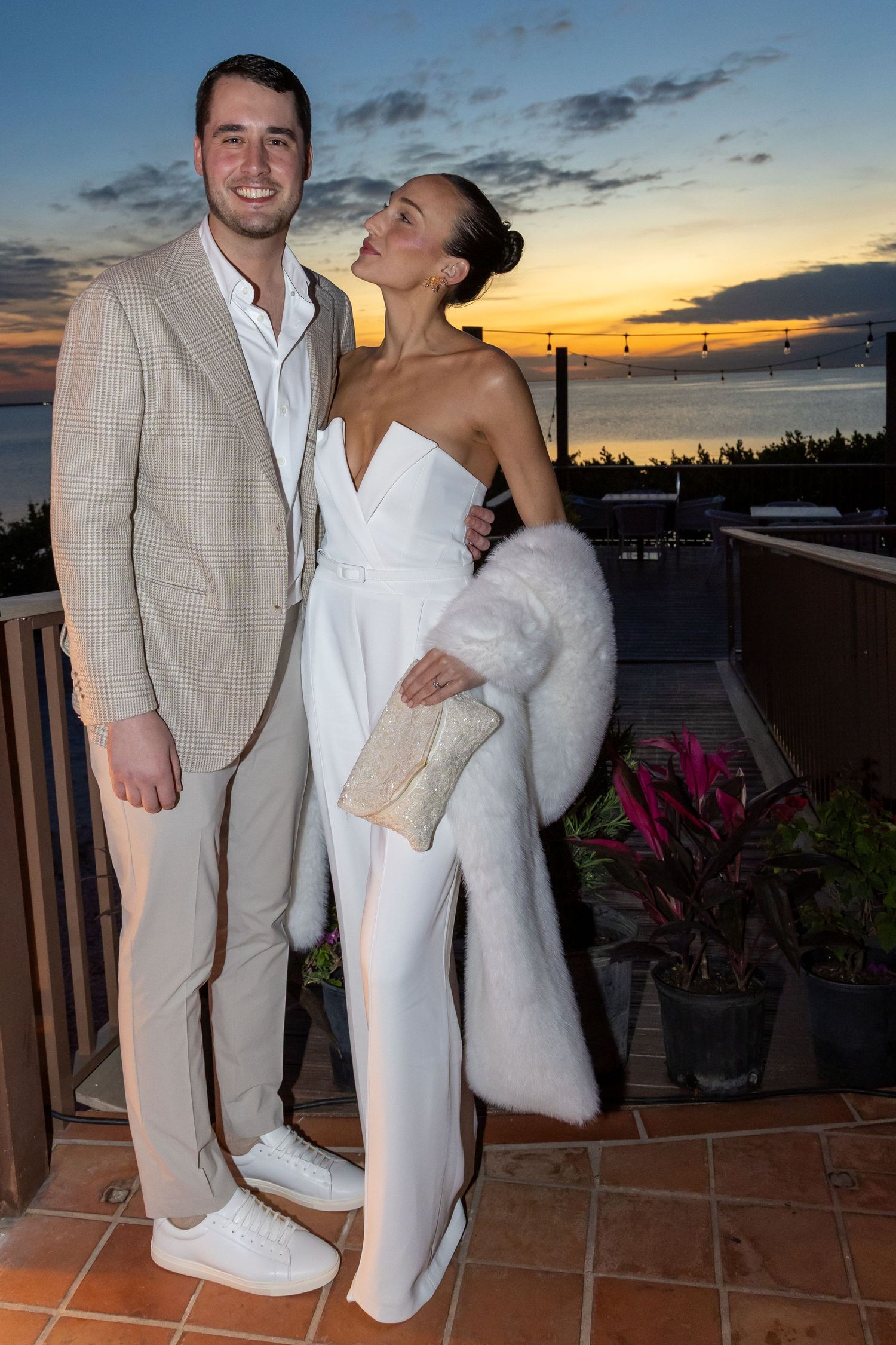 Elegant couple posing at sunset rooftop, man in tan suit, woman in white jumpsuit with fur stole.
