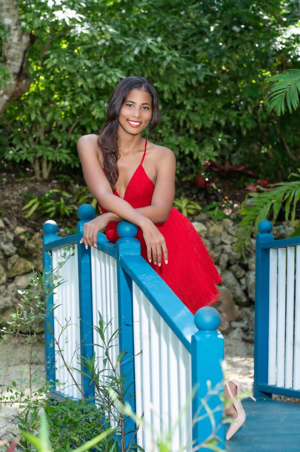 Young woman in red dress leaning on blue and white bridge railing surrounded by lush tropical greenery.