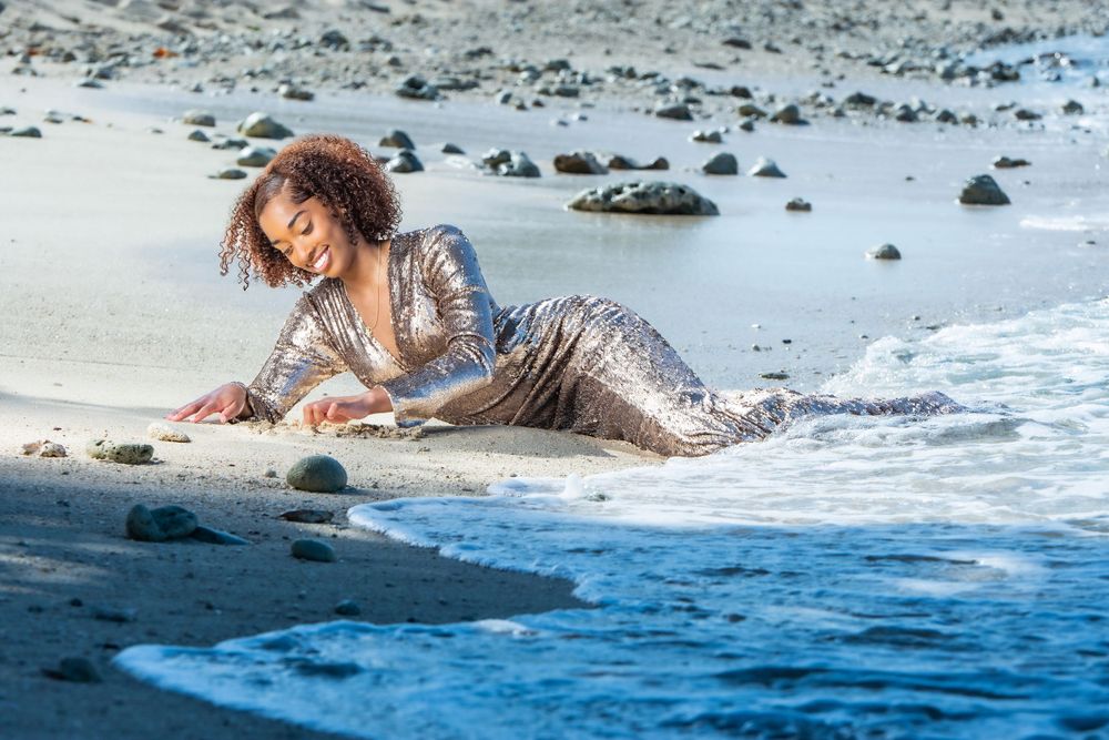 Woman in silver sequin gown poses gracefully on sandy beach with ocean waves at her feet.