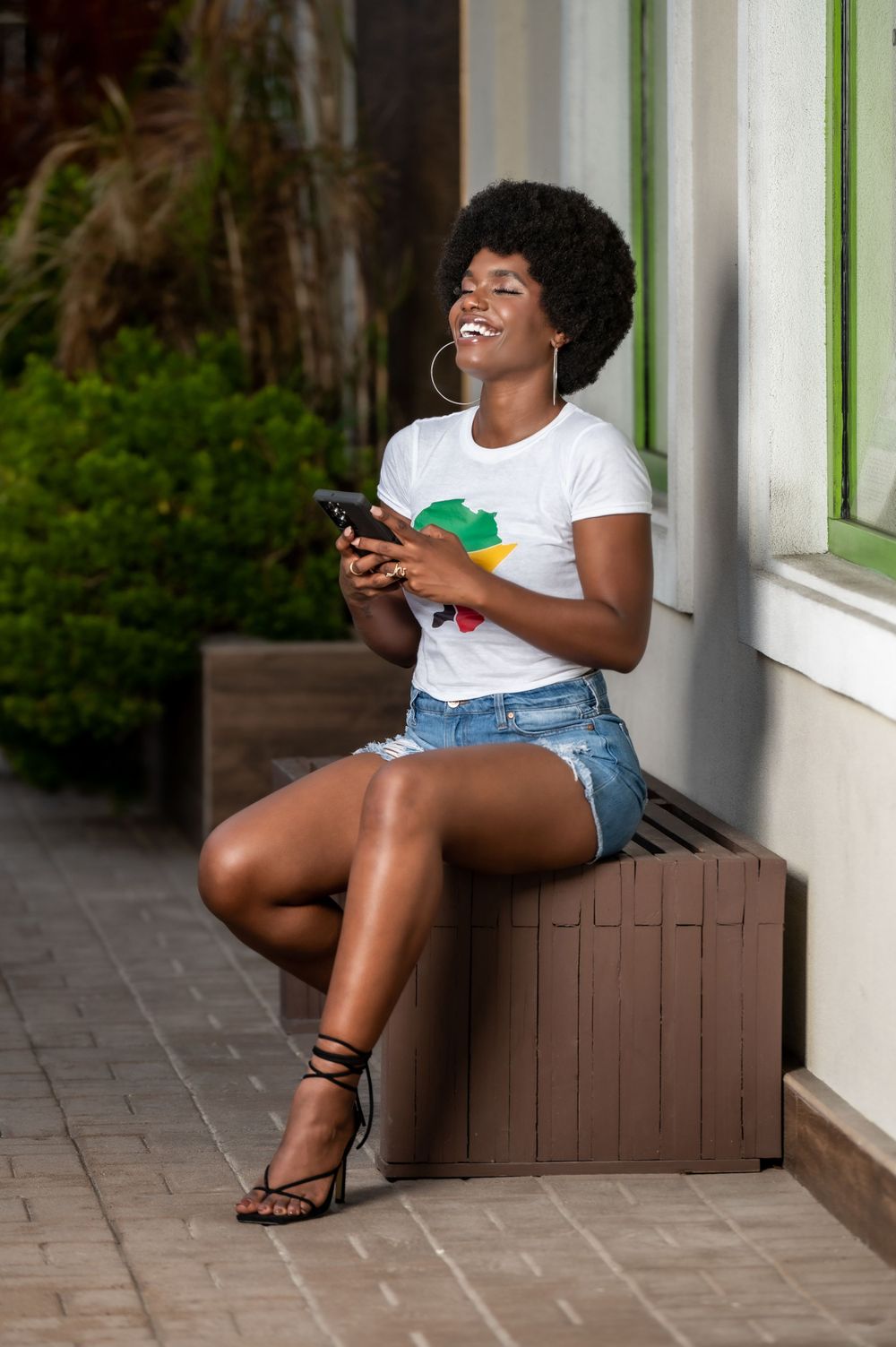 Smiling Black woman with afro hair sitting outdoors, wearing white graphic tee and denim shorts, holding smartphone.