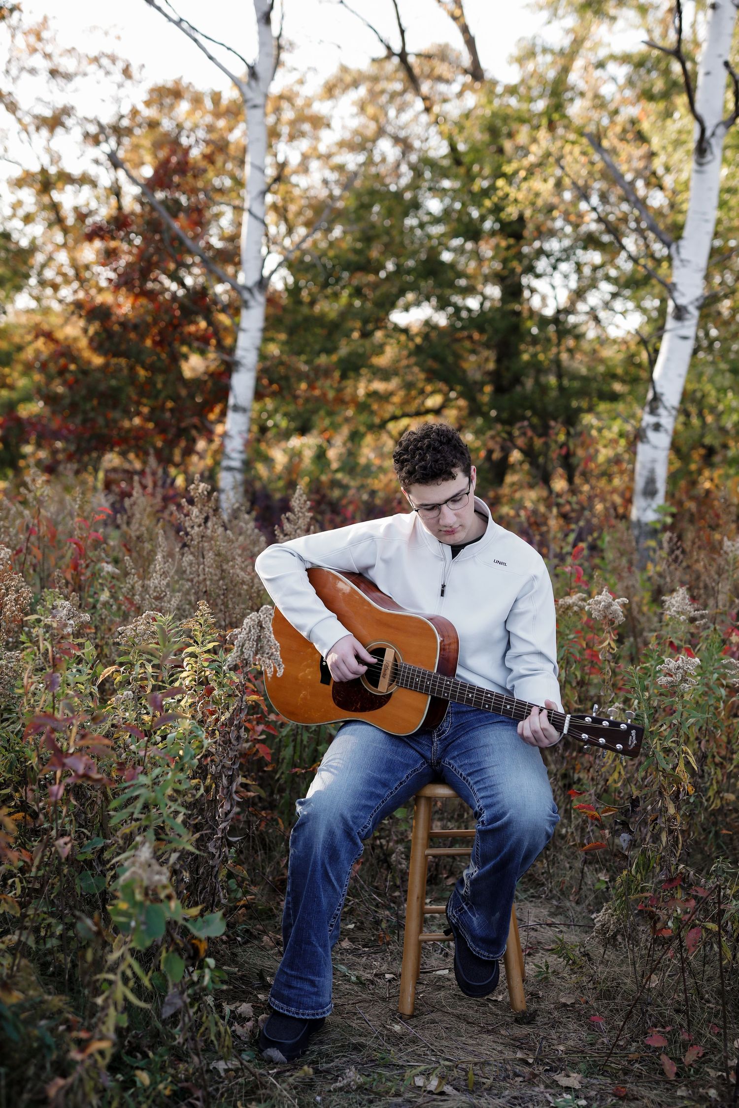 Young man playing acoustic guitar while seated outdoors in colorful autumn foliage setting.