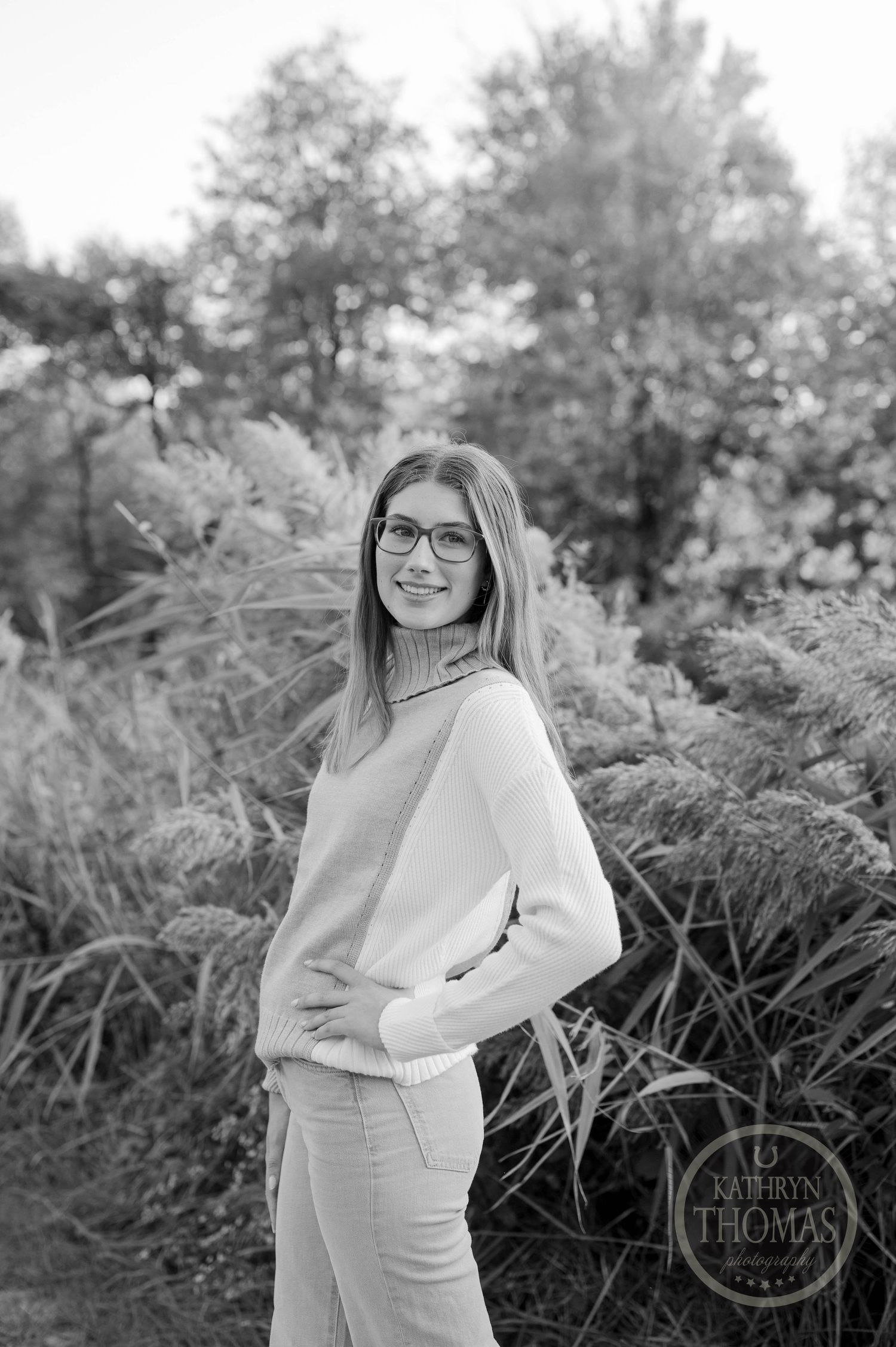 Black and white portrait of someone in glasses and sweater posing outdoors among natural elements.