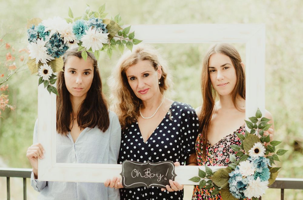 Three women in floral and polka dot dresses pose with decorative blue floral arrangements at an outdoor event.