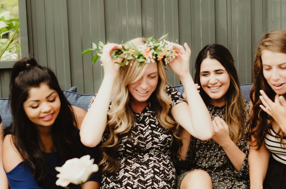 A group of friends in lace dresses laugh joyfully together while sitting outdoors during a summer gathering.