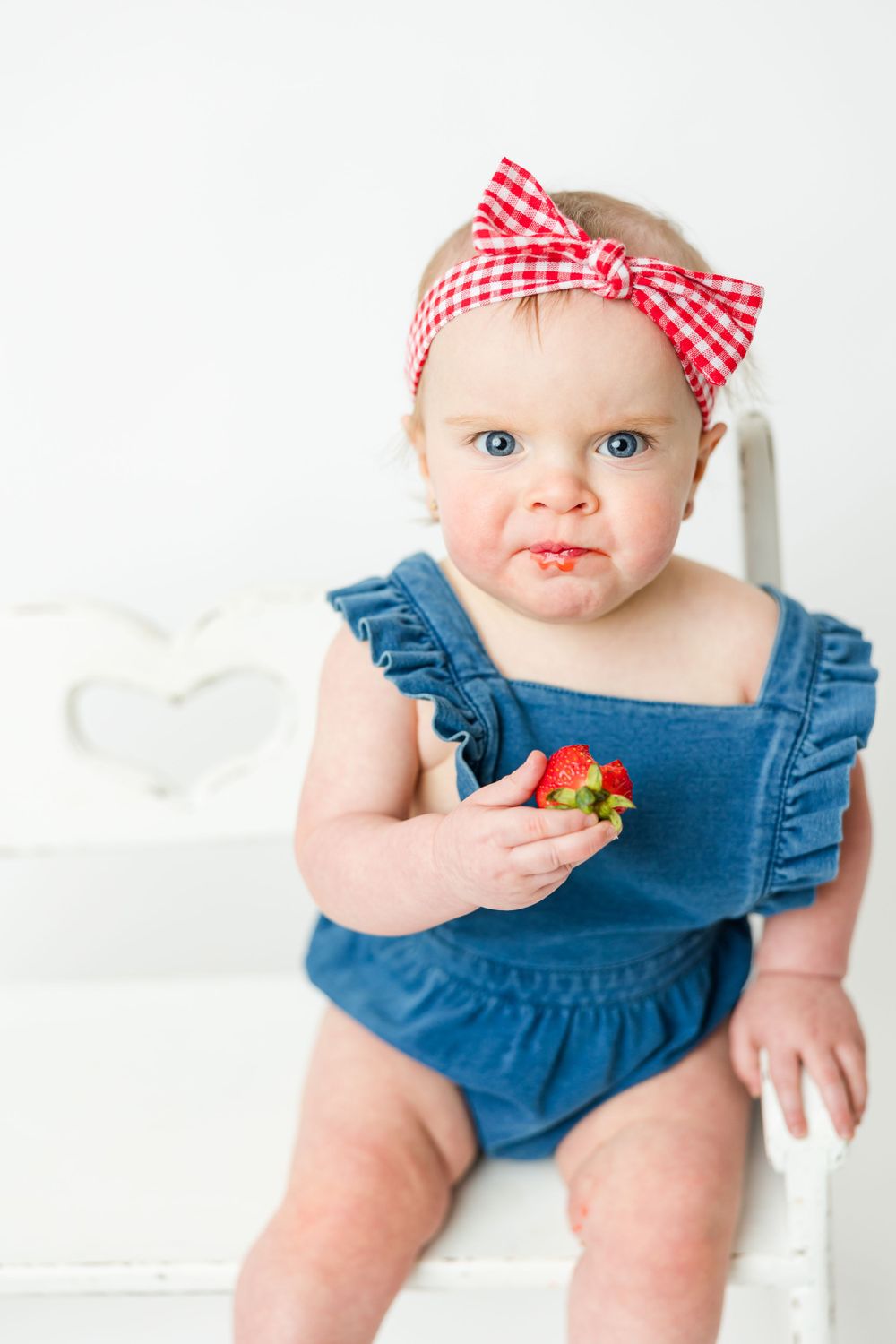 Baby in denim romper and red gingham bow headband holds strawberries while sitting on white chair.
