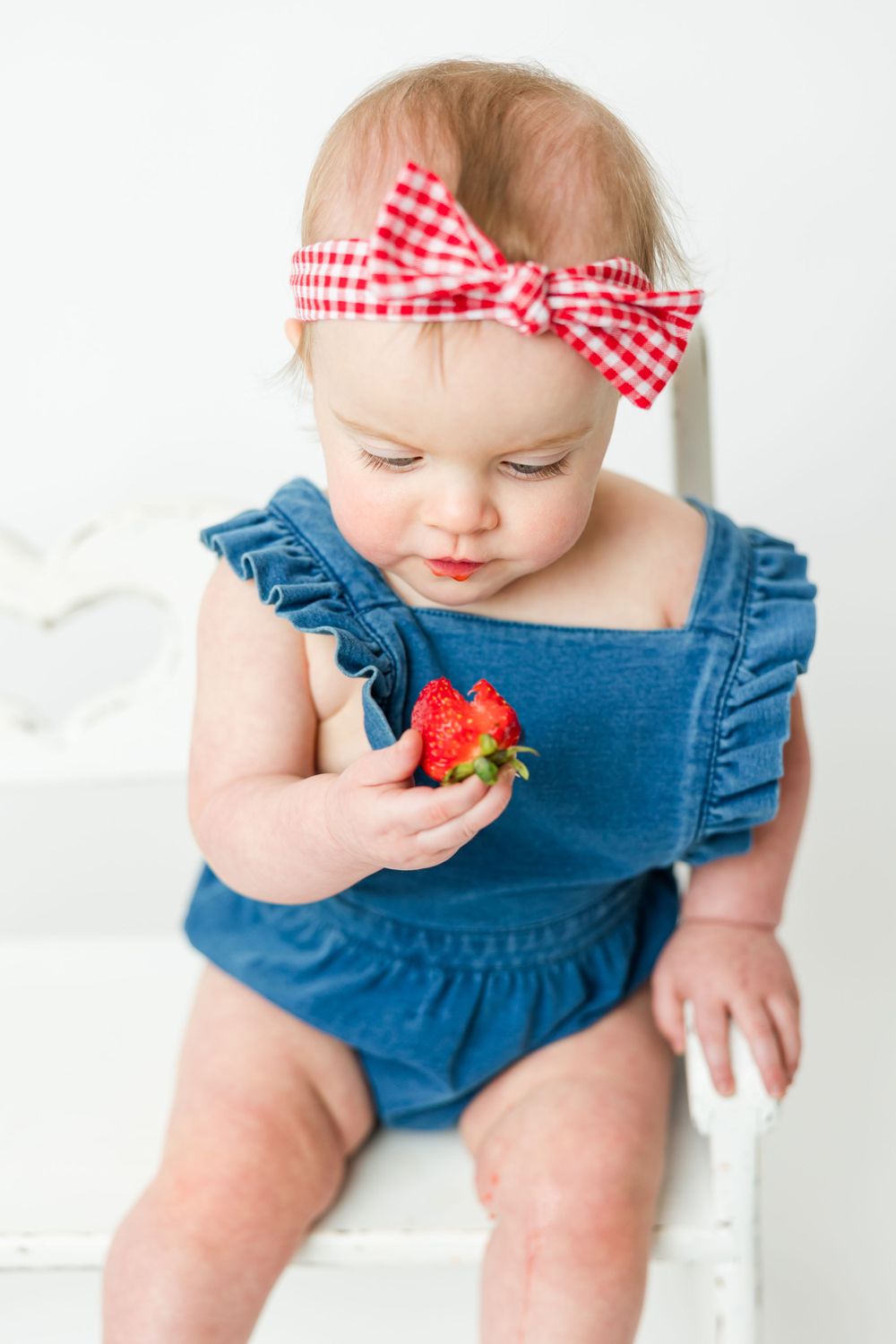 Sweet baby wearing denim romper and red gingham bow examines fresh strawberry while sitting on white chair.