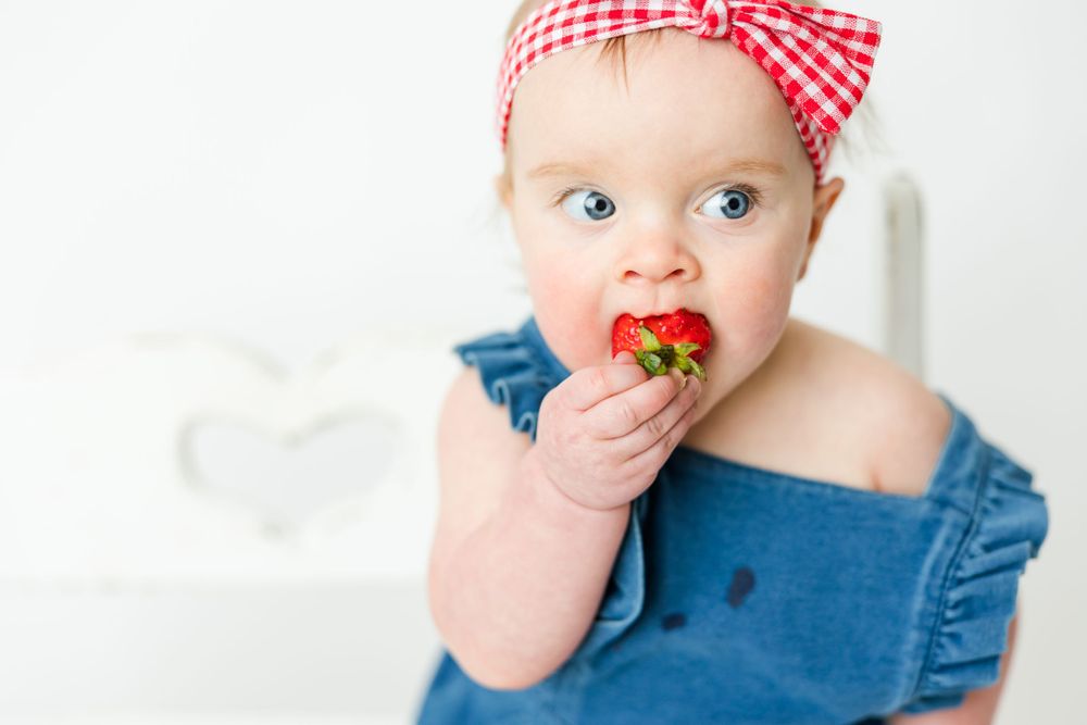 Baby wearing denim and red gingham headband enjoys eating fresh strawberry against white background.