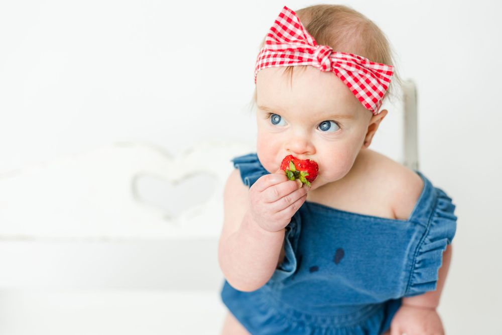 Sweet baby wearing denim and red checkered bow eats fresh strawberry against white background.