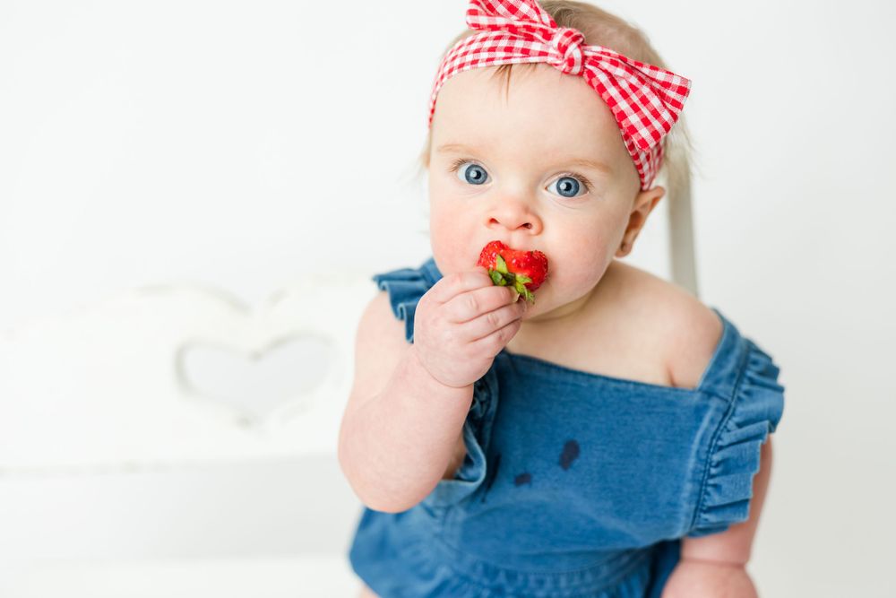 Baby wearing red checkered headband and denim romper sits with pacifier in white studio setting.