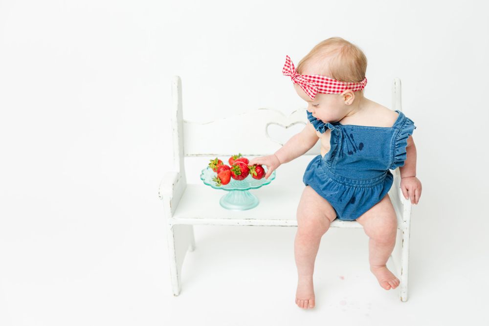 Baby in blue romper and headband sitting on white bench reaching for bowl of fresh tomatoes.