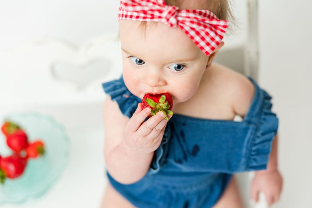Baby in denim overalls and gingham headband eating a fresh strawberry with a teal plate in background.