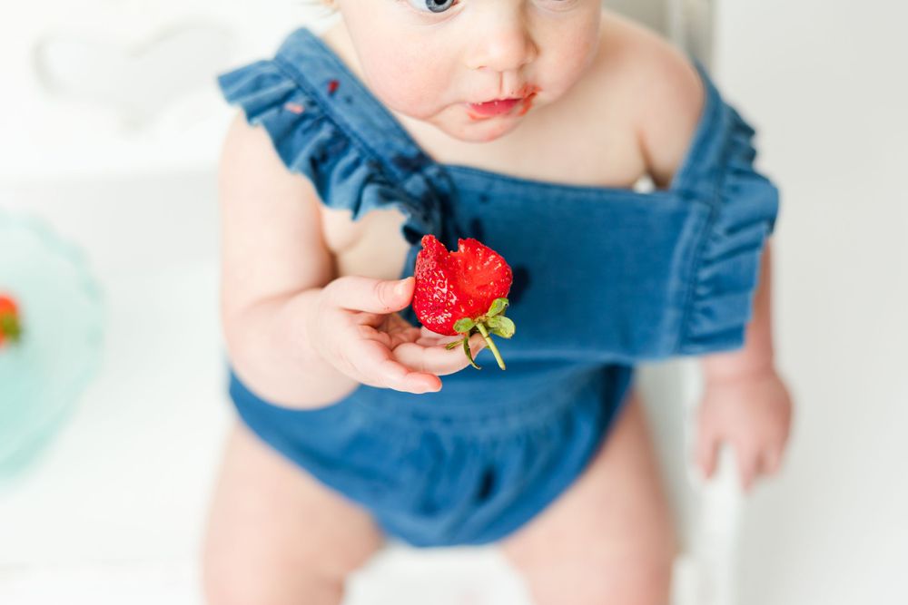 A small hand holds a fresh red strawberry while wearing a ruffled denim outfit.