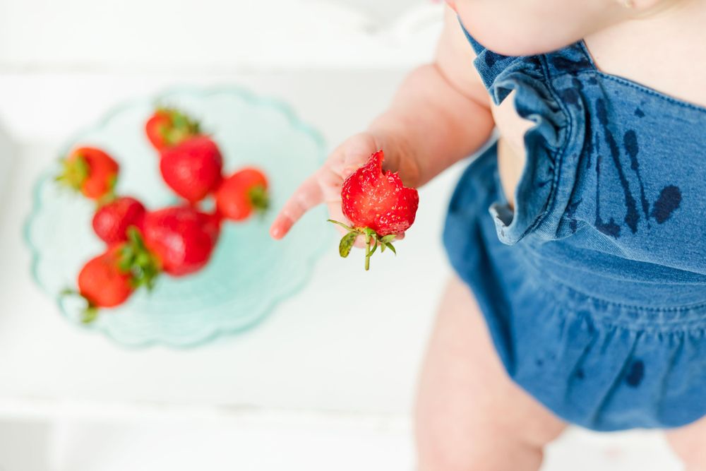 Fresh red strawberry being held in front of blue ceramic plate with more strawberries scattered on white surface.