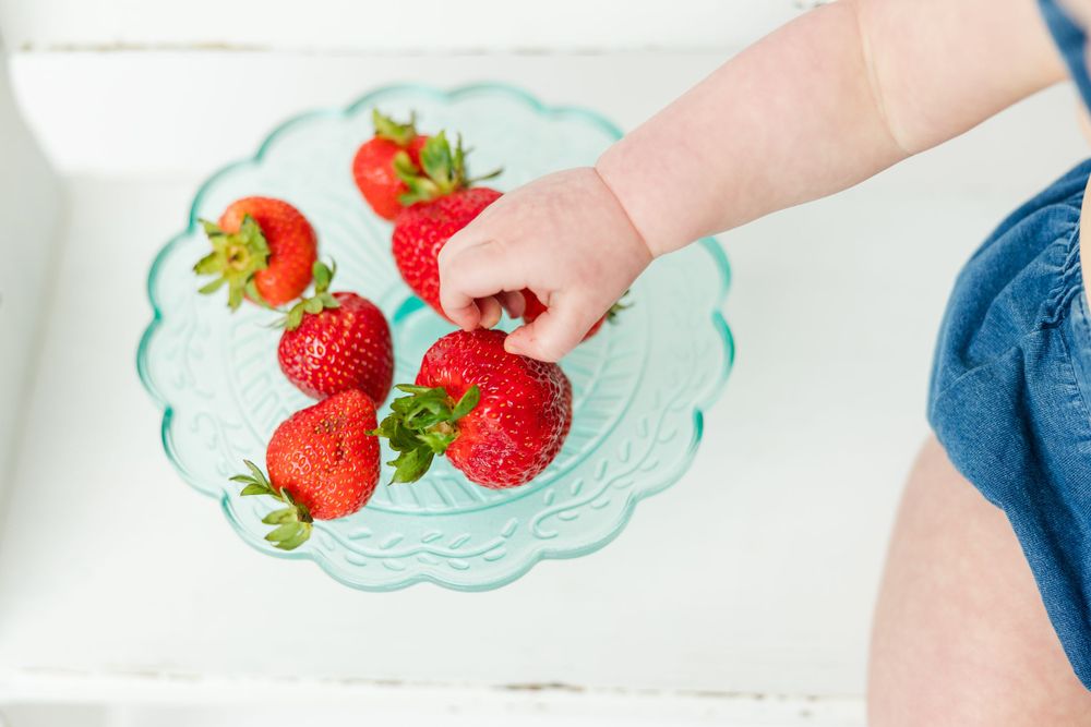 A small hand reaches for fresh red strawberries arranged on a decorative mint green glass plate.