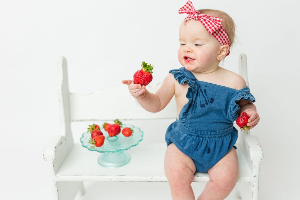 A small child in blue clothing sits with strawberries on a white chair near a turquoise plate.
