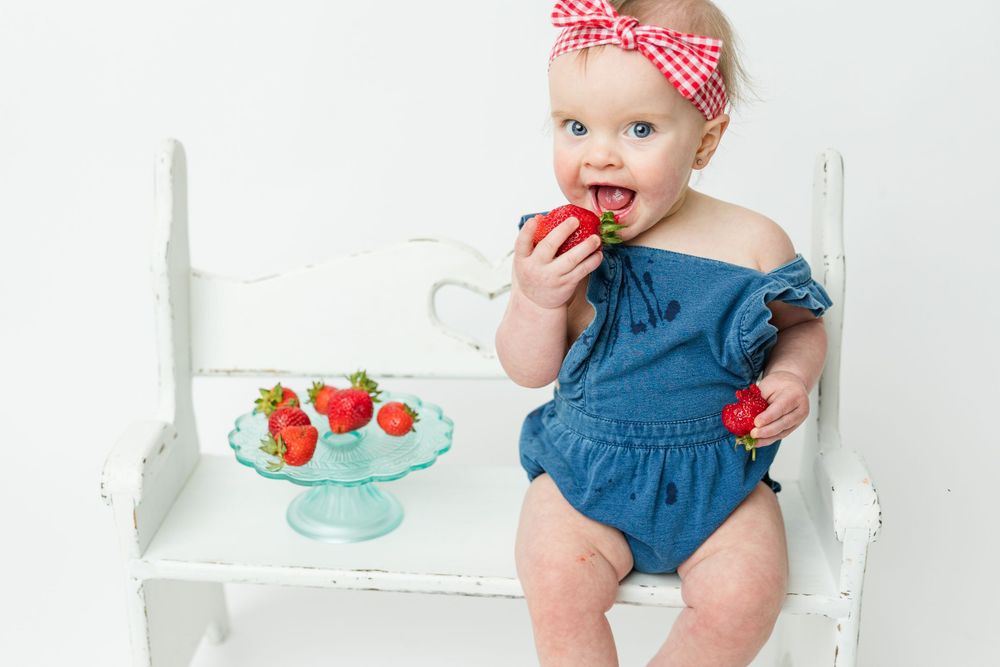 Small child in blue romper sitting on white bench eating fresh strawberries from turquoise glass stand.