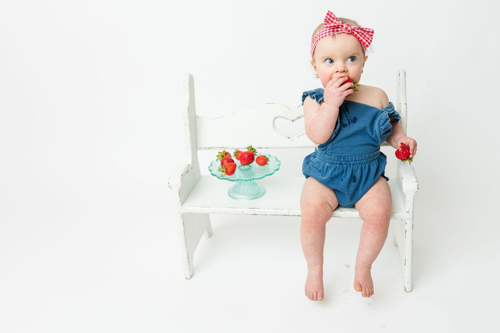 A small child in blue overalls sits on a white bench with strawberries and a decorative bow accessory.