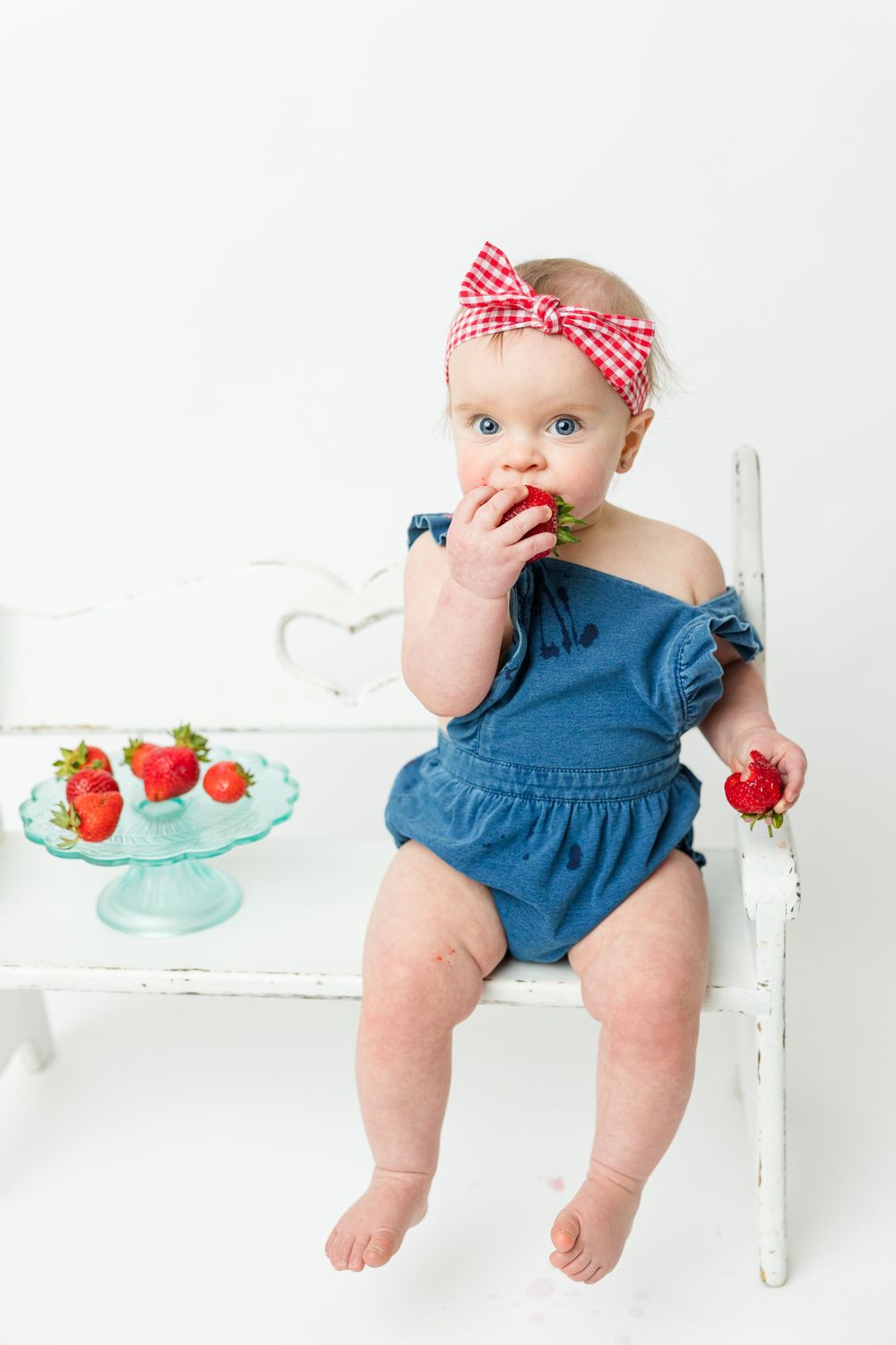 Baby in blue romper and red gingham headband enjoys fresh strawberries while sitting on white vintage chair.
