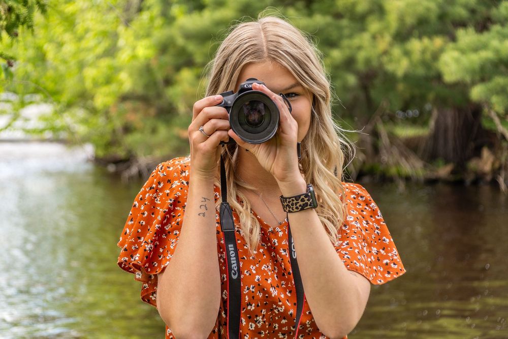 Someone wearing an orange floral dress takes photos with a professional camera near a river.